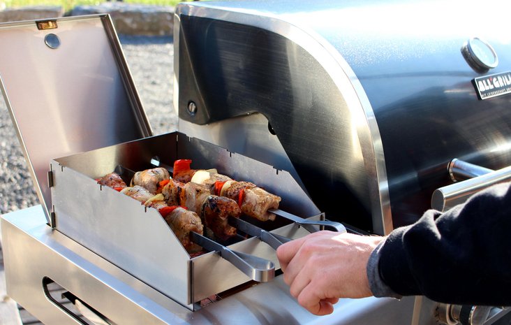 Schaschlik Hand hält Schaschlikspieße, die auf einem Grill in einer Auffangschale liegen und gegrillt werden.