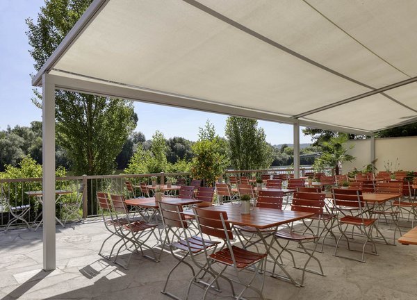 Biergarten mit Überdachung Überdachter Biergarten mit mehreren Tischen und roten Stühlen, umgeben von Bäumen und mit Blick auf einen Fluss.