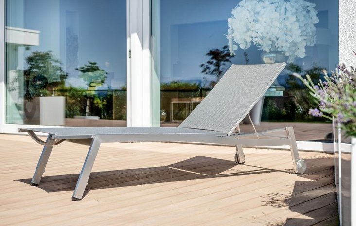 Terrasse mit Gartenliege Eine moderne, graue Gartenliege steht auf einer Holzterrasse vor großen Fensterscheiben.