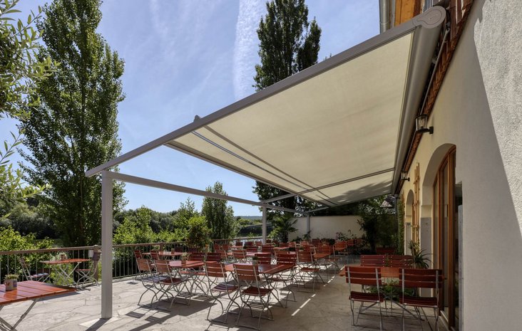 Biergarten mit Pergolamarkise Biergarten mit Pergolamarkise mit Holzstühlen und Tischen, umgeben von Bäumen und bepflanztem Geländer.