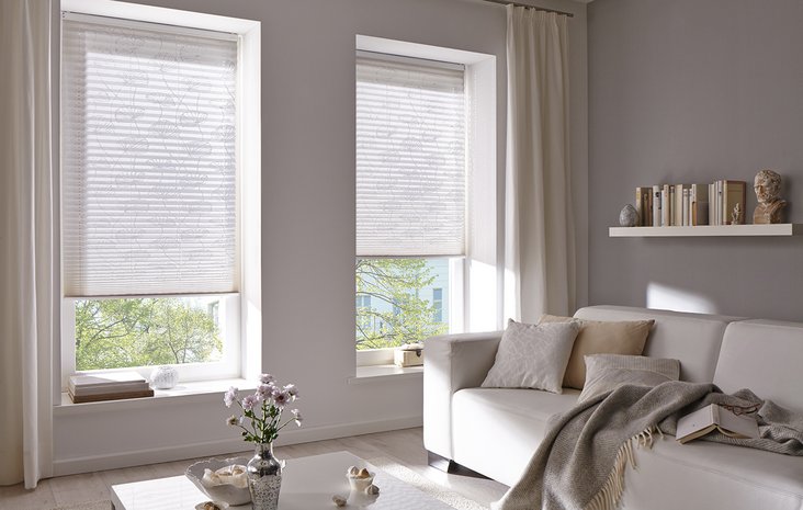 Fenster mit Kadeco Sonnenschutz Helles Wohnzimmer mit zwei großen Fenstern, die teils mit weißen Kadeco Jalousien bedeckt sind.