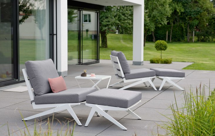 Stern Jackie Gartenliege Terrasse mit zwei modernen, weißen Stern Jackie Gartenliegen mit grauer Polsterung und dazu passenden Hockern vor einem Glasfenster.
